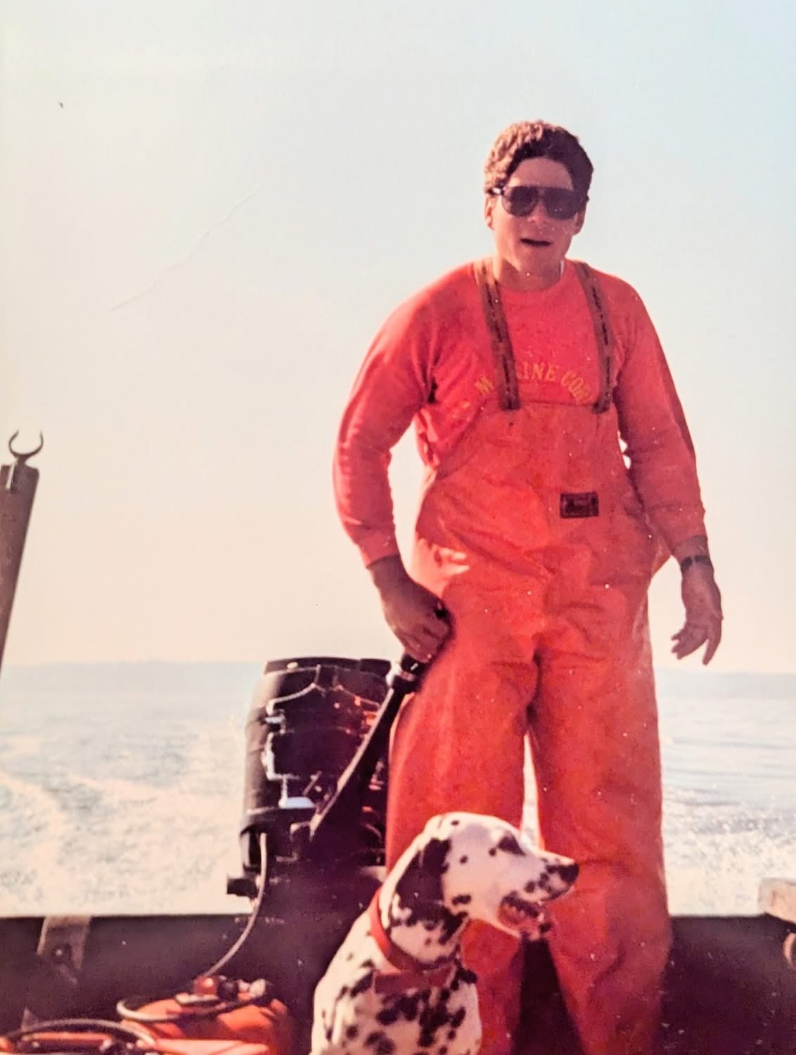 Long Island bayman in orange foul-weather bibs aboard his boat with a dalmatian at his side